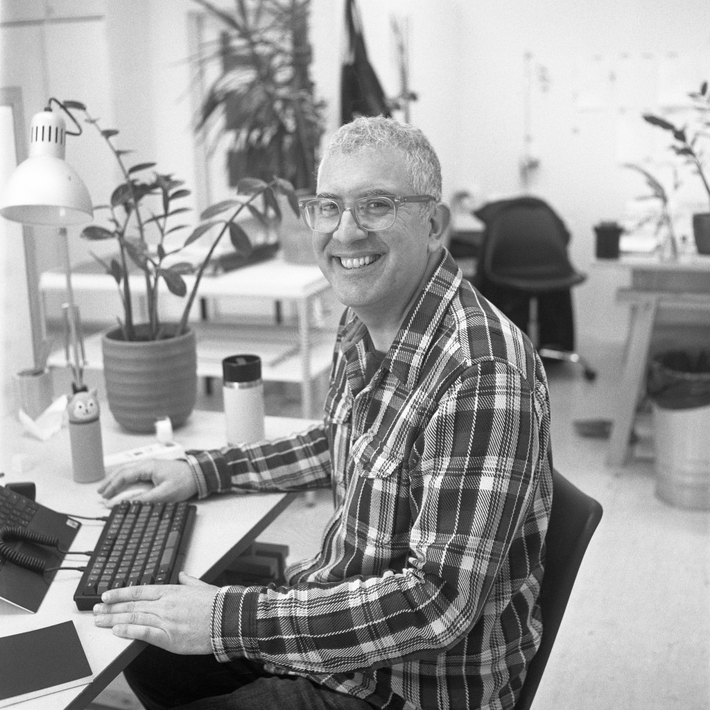 A smiling man sits at a desk with his hands on a keyboard.